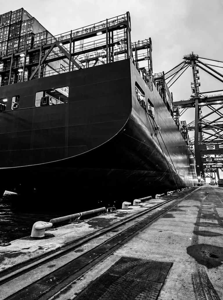 Black-and-white photograph of a ship moored beside a quay, with scaffolding and cranes behind it